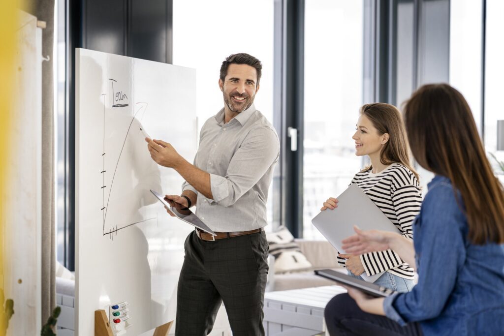 Smiling businessman with digital tablet explaining market development fund projections to female colleagues at office using a whiteboard.