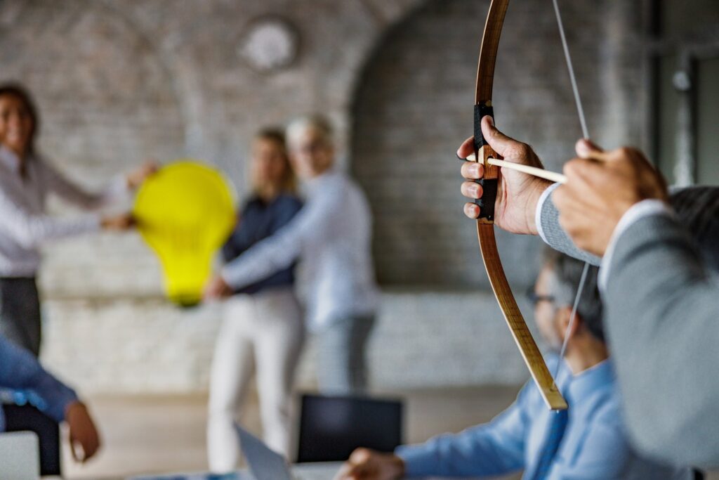 Close up of unrecognizable male entrepreneur shooting at target with bow and arrow in the office. There are people in the background.