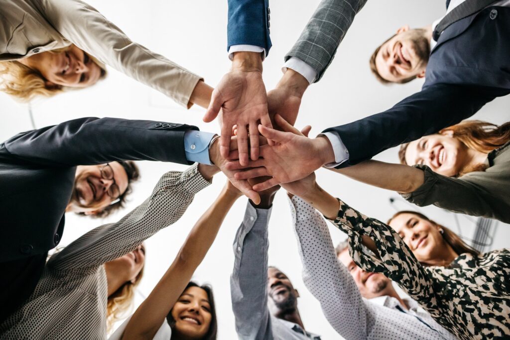 A worm's eye view close-up of female and male colleagues in a business stacking their hands together. They are cheering and laughing. All wear fashionable business clothes. Horizontal daylight indoor photo.